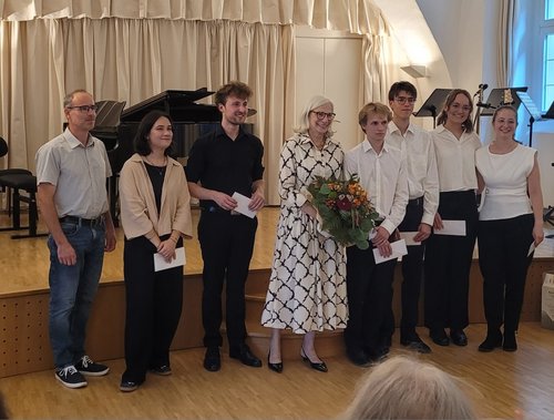 Gruppenfoto der Mitwirkenden nach einem Konzert auf der Bühne eines Konzertsaals mit applaudierendem Publikum.