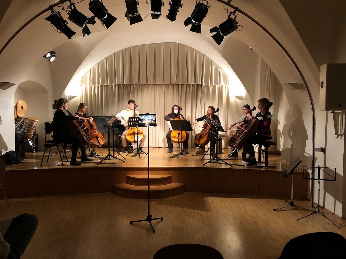 Acht Cellist*innen sitzen auf der Bühne eines Konzertsaals und musizieren