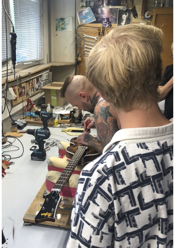 Ein blonder Schüler ist von hinten zu sehen, wie er einem Mitarbeiter der Firma Höfner bei der Arbeit an einem Instrument zusieht.