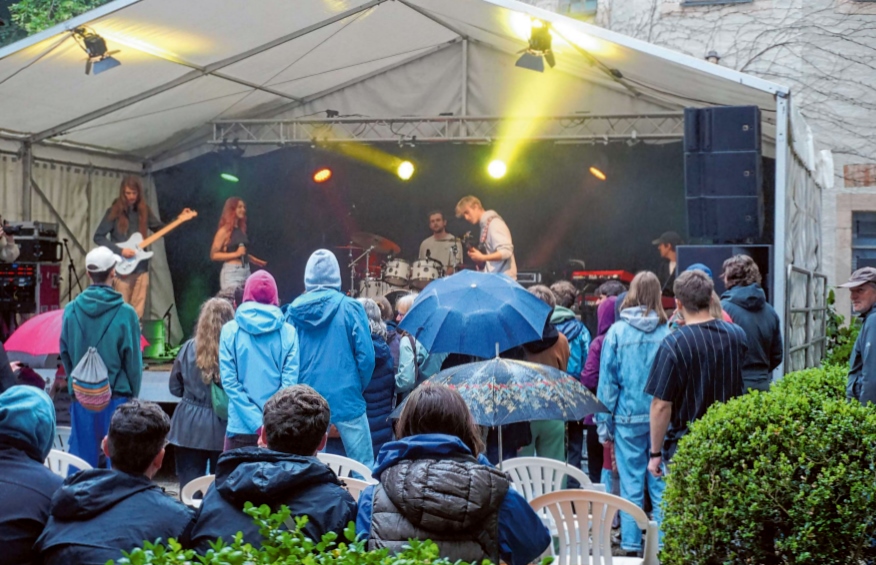 Blick vom leicht verrregneten Zuschauer-Bereich auf die Bühne, wo eine Band in Aktion ist.